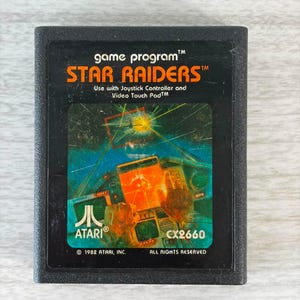 May include: A black Atari Star Raiders game cartridge for the 2600 console. The label features orange text reading Star Raiders, game program, and the Atari logo. The central illustration depicts a cockpit view of a space battle with laser fire.
