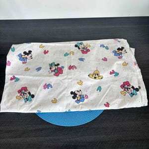 Könnte beinhalten: Weißes Spannbettlaken mit einem sich wiederholenden Muster von Mickey und Minnie Mouse in verschiedenen Posen sowie bunten Formen. Das Laken ist gefaltet und liegt auf einem blauen Platzset.