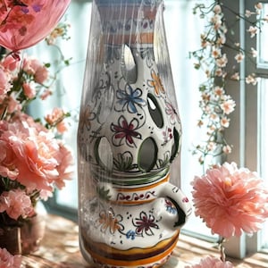 Puede incluir: Un jarrón de cerámica blanco con asa, adornado con diseños florales coloridos. El jarrón tiene aberturas recortadas y está envuelto en plástico transparente. El jarrón está decorado con detalles en naranja, verde, azul y rojo.