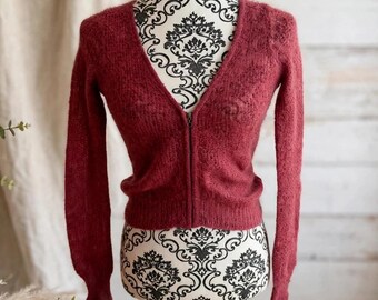 May include: A deep red, long-sleeved cardigan with a V-neckline and a full-length front zipper closure. The garment is crafted from a textured, fuzzy knit fabric and features ribbed cuffs and a ribbed hemline for a fitted silhouette.