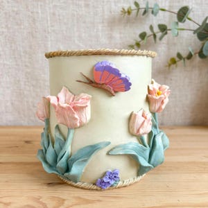 May include: A cylindrical cream-coloured ceramic vase featuring raised relief decorations of pink tulips with light blue leaves, a purple and orange butterfly, and small purple flowers, finished with a braided rope-style trim at the top and base.