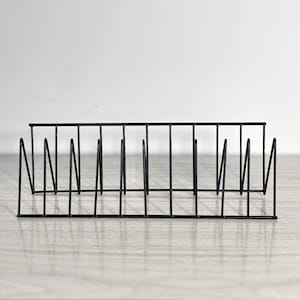 Puede incluir: Un estante de alambre negro con múltiples divisores verticales. El estante rectangular está diseñado para sostener objetos en posición vertical. El metal es delgado y el diseño es minimalista. Se encuentra sobre una superficie clara.