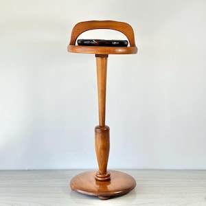 Può includere: Un supporto in legno con base rotonda, colonna centrale sottile e parte superiore circolare con manico curvo. Il supporto è in legno marrone chiaro. La parte superiore contiene un oggetto scuro, forse di vetro. Design vintage.