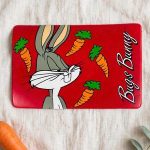 May include: A rectangular red tray featuring a graphic of the cartoon character Bugs Bunny with long ears and white whiskers. Three orange carrots with green tops are positioned around the character. The text Bugs Bunny is printed in white.