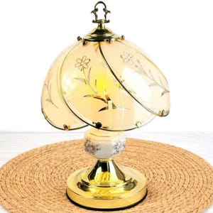Peut inclure: Une lampe de table de style vintage avec une base et des accents dorés. L'abat-jour en verre de couleur crème est orné d'un motif floral. La base présente un motif floral décoratif. La lampe est posée sur un tapis rond tissé.