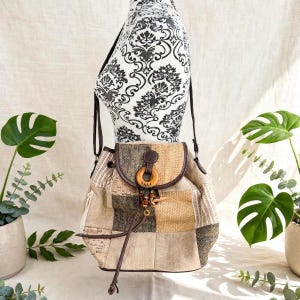 May include: A patchwork shoulder bag featuring various textured fabric panels in shades of beige, tan, brown, and black. The bag has a flap closure secured with a dark brown tab and a circular wooden ring accent with dangling wooden beads.
