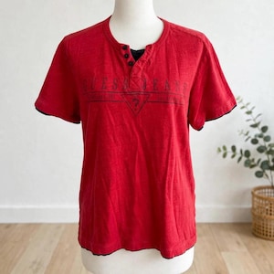 May include: A red short-sleeve henley shirt featuring a black three-button placket and a black graphic print on the chest that reads GUESS JEANS with a triangle logo containing a question mark. The shirt has a black inner lining visible at the neck.