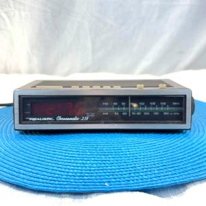May include: A vintage Realistic Chronomatic-238 digital clock radio. The clock displays red digital numbers and has an AM/FM radio dial. The face is silver and black, with the brand name and model number printed on the front.