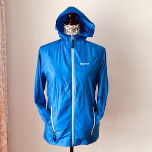 Peut inclure: Veste à capuche bleu vif avec une fermeture éclair blanche et le nom de la marque "Marmot" imprimé sur la poitrine. La veste a une capuche, deux poches avant et une bordure bleu clair. La veste est présentée sur un mannequin.