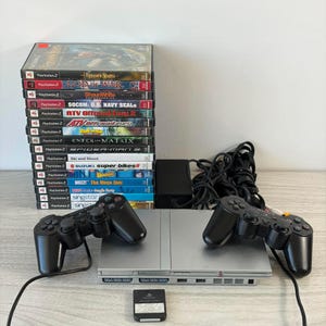 May include: A silver PlayStation 2 console with two black controllers, a memory card, and a stack of game cases. Games include "God of War," "Spider-Man," and "Buzz! The Mega Quiz."