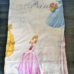 Puede incluir: Sábana blanca con ilustraciones de princesas de Disney con sus vestidos. La sábana tiene un diseño de guión azul claro con la palabra "Princess" y frases como "Dear Diary" y "I'm going to the Ball!"