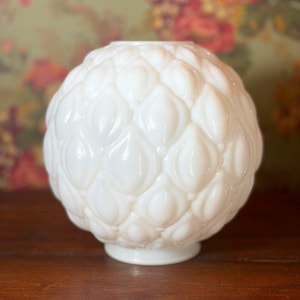 May include: A white, spherical glass lamp shade with a textured, petal-like pattern. The shade has a rounded shape and sits on a small, circular base. The background is a blurred floral pattern.