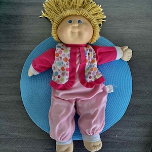 Può includere: Una bambola vintage con un viso rotondo, occhi blu e capelli di filo giallo. La bambola indossa un completo rosa con un gilet fantasia ed è appoggiata su un tappetino circolare blu. Le braccia della bambola sono tese.