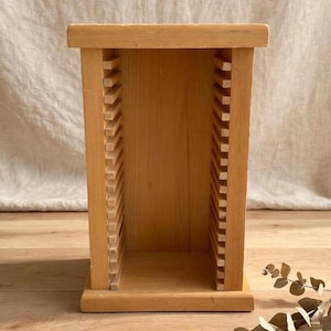Peut inclure: Un support de rangement en bois clair doté d'un cadre rectangulaire avec un dessus et une base plats. Les côtés intérieurs sont garnis de nombreuses lattes de bois horizontales régulièrement espacées pour maintenir des objets à la verticale.