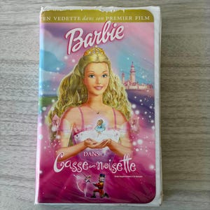 May include: A Barbie in the Nutcracker VHS tape featuring a blonde character wearing a gold crown and a white dress with pink straps. The cover displays the text Barbie and Casse-noisette, with a small fairy, a castle, and a ballet scene below.