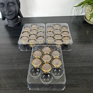 Peut inclure: Des boutons de meubles avec une base dorée et un dessus circulaire de style antique, disposés dans des plateaux en plastique transparent. L'image montre également une statue de Bouddha noire et une plante en pot.