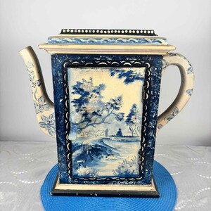 May include: A decorative, rectangular teapot with a blue and white landscape painting on the front. The teapot has a cream-colored handle and spout with floral designs. The lid is cream-colored with a black border and white dots.