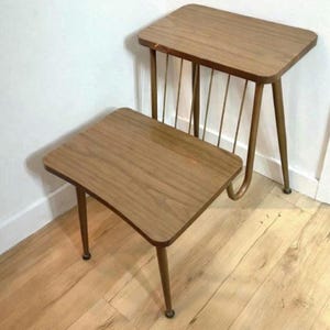 Puede incluir: Dos mesas auxiliares de madera vintage con un acabado marrón cálido. Las mesas tienen una parte superior rectangular y patas delgadas e inclinadas. Una mesa presenta un diseño de revistero con varillas metálicas verticales. Las mesas están sobre un suelo de madera.