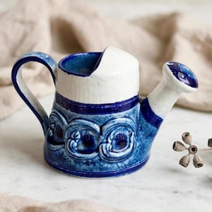 Peut inclure: Un arrosoir en céramique avec une partie supérieure blanche et une partie inférieure bleu foncé ornée de motifs circulaires en relief. La poignée et le bec sont intégrés au corps, avec une tête de bec blanche et une poignée bleue à accents blancs.