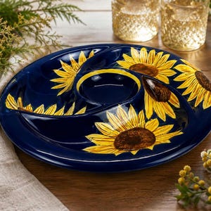 May include: A round, dark blue ceramic serving platter featuring a central circular compartment surrounded by three curved sections. The surface is decorated with large, vibrant yellow sunflowers with brown centres painted across the navy blue base.
