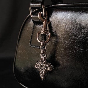 K&ouml;nnte beinhalten: Eine schwarze Ledertasche mit einer silbernen Kette und einem dekorativen Kreuzanh&auml;nger.