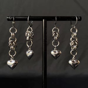 May include: Four silver heart-shaped dangle earrings with a chain link design. The earrings are displayed on a black stand.