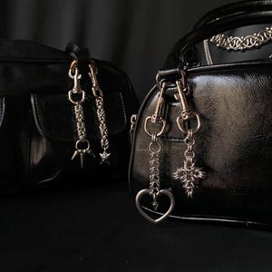 K&ouml;nnte beinhalten: Zwei schwarze Ledertaschen mit silbernen Ketten- und Anh&auml;nger-Akzenten. Eine Tasche hat einen Stern- und Dornenanh&auml;nger, die andere einen Herz- und Kreuzanh&auml;nger.