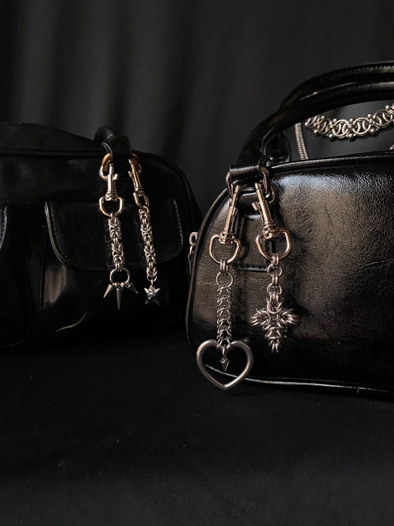 K&ouml;nnte beinhalten: Zwei schwarze Ledertaschen mit silbernen Ketten- und Anh&auml;nger-Akzenten. Eine Tasche hat einen Stern- und Dornenanh&auml;nger, die andere einen Herz- und Kreuzanh&auml;nger.