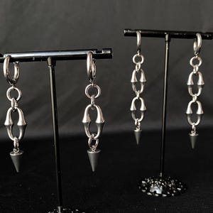 May include: Two pairs of silver-toned earrings on display. Each earring features a hoop at the top, connected to a series of linked, curved metal pieces, and a dark gray, cone-shaped pendant at the bottom. The earrings are displayed on black stands.