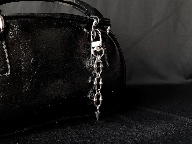 May include: A black, glossy handbag with a silver-toned chain and a pointed charm. The chain is attached to the handle of the bag. The background is a dark, solid color.