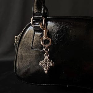 K&ouml;nnte beinhalten: Eine schwarze Ledertasche mit einer silbernen Kette und einem silbernen Anh&auml;nger in Form eines Kreuzes.