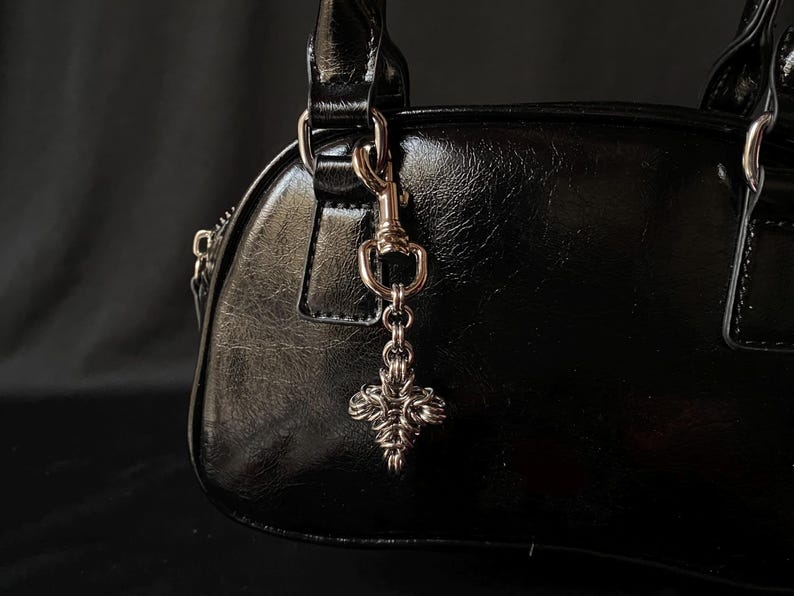 K&ouml;nnte beinhalten: Eine schwarze Ledertasche mit einer silbernen Kette und einem silbernen Anh&auml;nger in Form eines Kreuzes.
