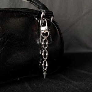 May include: A black, shiny handbag with a silver-colored chain and a pointed charm. The chain is attached to the bag with a clasp. The bag is on a dark background.