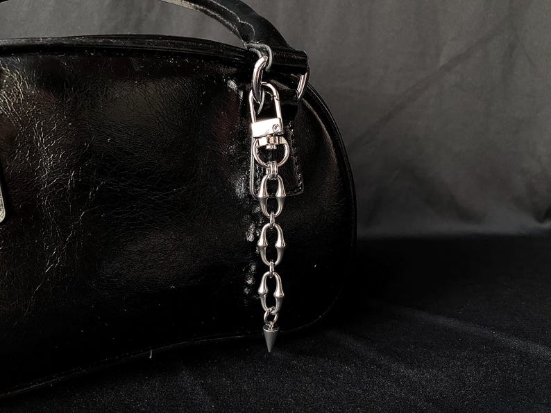 May include: A black, shiny handbag with a silver-colored chain and a pointed charm. The chain is attached to the bag with a clasp. The bag is on a dark background.