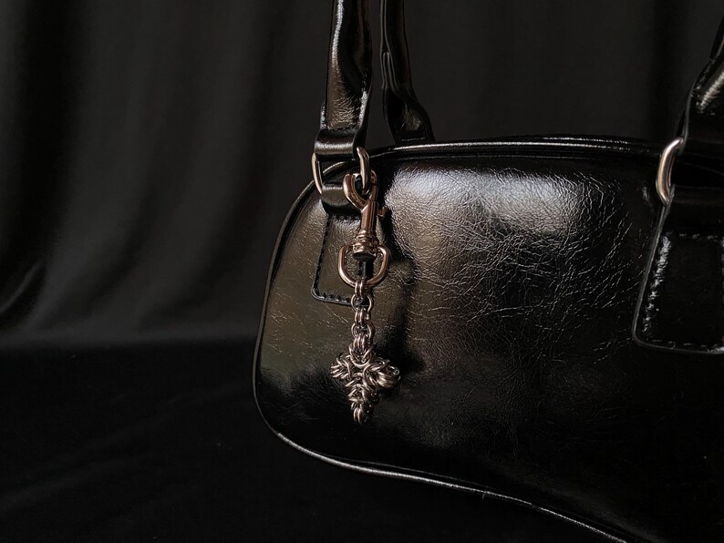 K&ouml;nnte beinhalten: Eine schwarze Ledertasche mit einer silbernen Kette und einem dekorativen Kreuzanh&auml;nger.