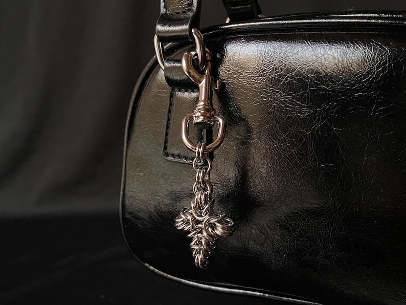 K&ouml;nnte beinhalten: Eine schwarze Ledertasche mit einem silbernen Anh&auml;nger in Form eines Kreuzes an einer Kette.