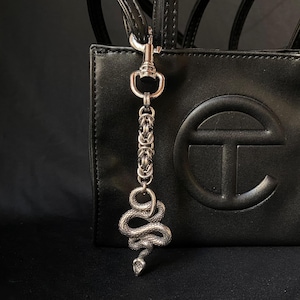 May include: A silver snake charm with a chain and clasp, suspended from a black handbag. The snake is intricately detailed with scales and a coiled form. The handbag features a large embossed logo.