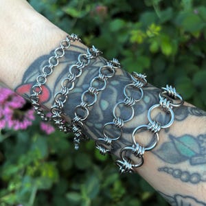May include: Five silver chain bracelets with varying link designs. The bracelets are worn on an arm with tattoos. The bracelets are made of shiny metal and are displayed against a blurred green and pink background.