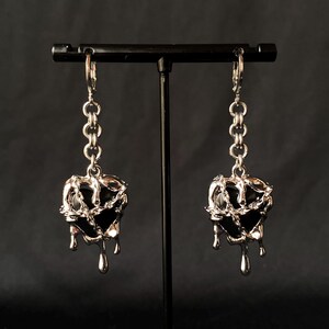 May include: A pair of silver earrings with black heart-shaped charms. The charms are dripping with silver, giving the appearance of melting.