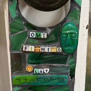 May include: A mosaic door plate with a dark metal doorknob. The plate is green and features the words "OUT FISHING" and coordinates. A green face is also part of the design.