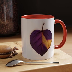 Puede incluir: Taza de cerámica blanca con interior y asa rojos. La taza presenta un diseño de hoja morada y dorada. Una cuchara plateada, un macaron y granos de café están sobre una superficie de madera.