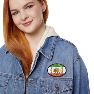 Può includere: Toppa ovale con l'emblema della bandiera iraniana, un leone e un sole su sfondo bianco, bordata da strisce verdi e rosse. La toppa è applicata su una giacca di jeans.