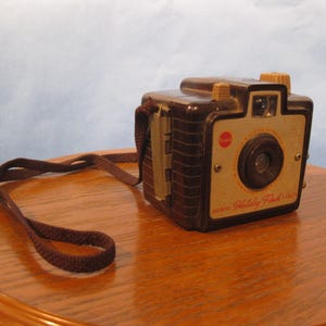 May include: Vintage Kodak Holiday Flash camera. The camera is brown and tan with a brown strap. The camera has a lens, flash, and a red circle on the front. The camera is sitting on a wooden table.