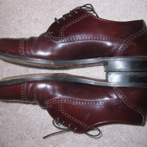 May include: A pair of brown leather lace-up shoes with a brogue design. The shoes have a dark brown sole and a pointed toe.