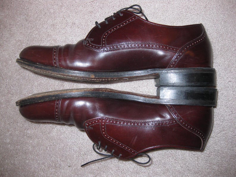 May include: A pair of brown leather lace-up shoes with a brogue design. The shoes have a dark brown sole and a pointed toe.