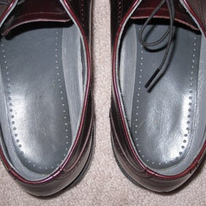 May include: A pair of burgundy leather shoes with black laces. The shoes have a gray insole with a dotted pattern.