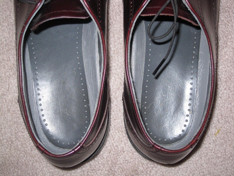 May include: A pair of burgundy leather shoes with black laces. The shoes have a gray insole with a dotted pattern.