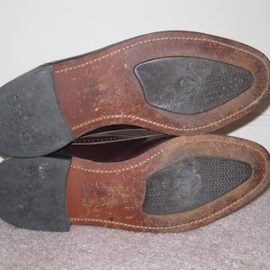 May include: Close-up of the soles of a pair of brown leather shoes. The shoes have a brown leather upper and a black rubber sole.