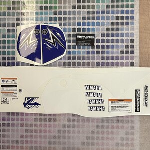 Peut inclure: Un kit graphique de moto Yamaha blanc et bleu avec le texte "YZ inger" et "BMCO DESIGN.COM". Le kit comprend un graphique d'aile avant, un graphique de panneau latéral et un graphique d'aile arrière. Les graphiques sont imprimés sur un matériau en vinyle transparent.