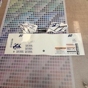 Peut inclure: Un kit graphique de moto Yamaha blanc avec des accents bleus et argentés. Le kit comprend un logo Yamaha, le numéro 80 et les lettres PW. Le kit est conçu pour une moto Yamaha.